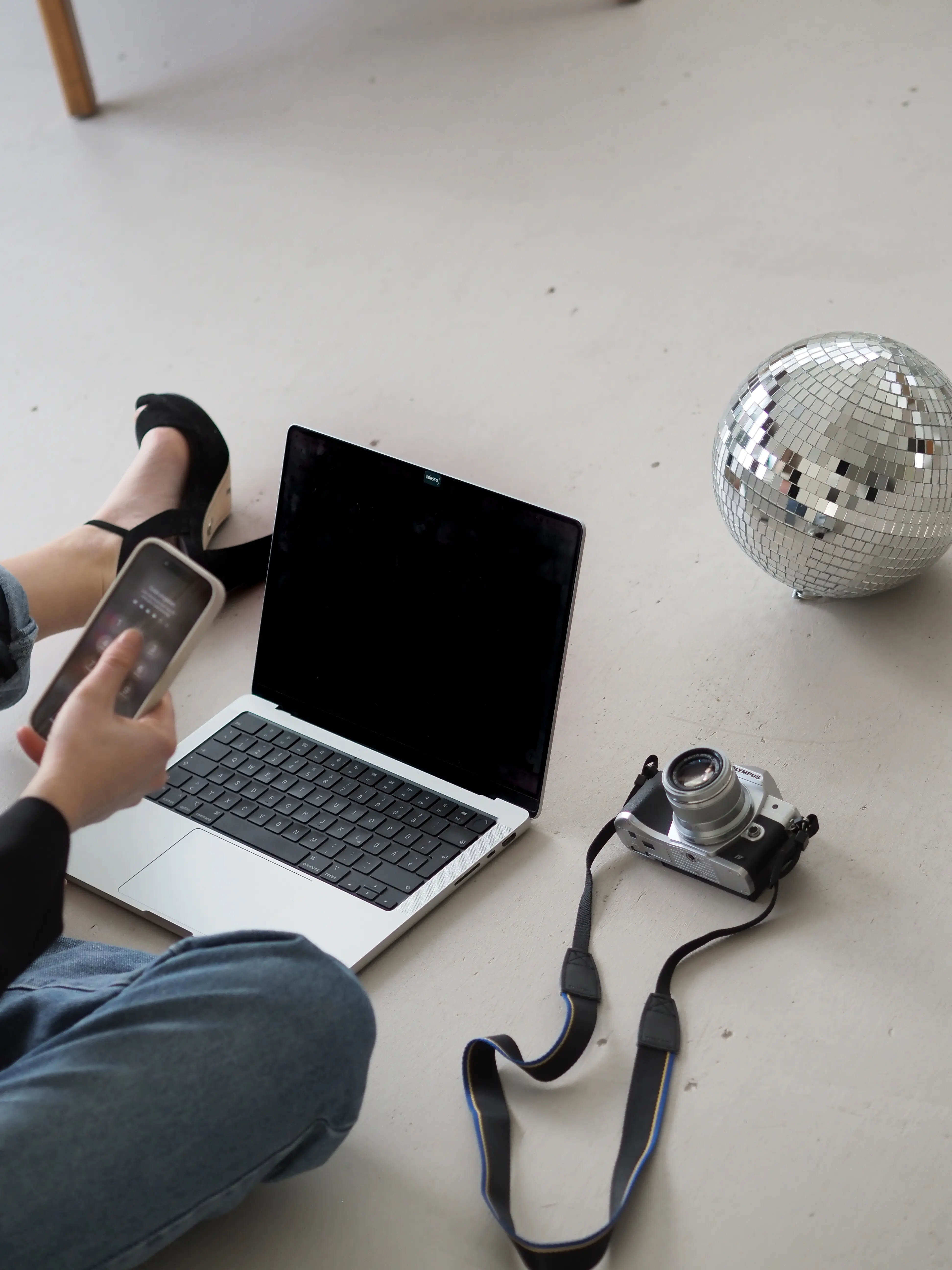 Eine Person sitzt auf dem Boden mit einem Smartphone in der Hand, neben einem Laptop mit schwarzem Bildschirm. Auf dem Boden liegen eine silberne Kamera und eine Diskokugel, die den Raum widerspiegelt. Die Szene symbolisiert Kreativität und bold ideas, mit einer Mischung aus Technologie und stilvollen Accessoires.