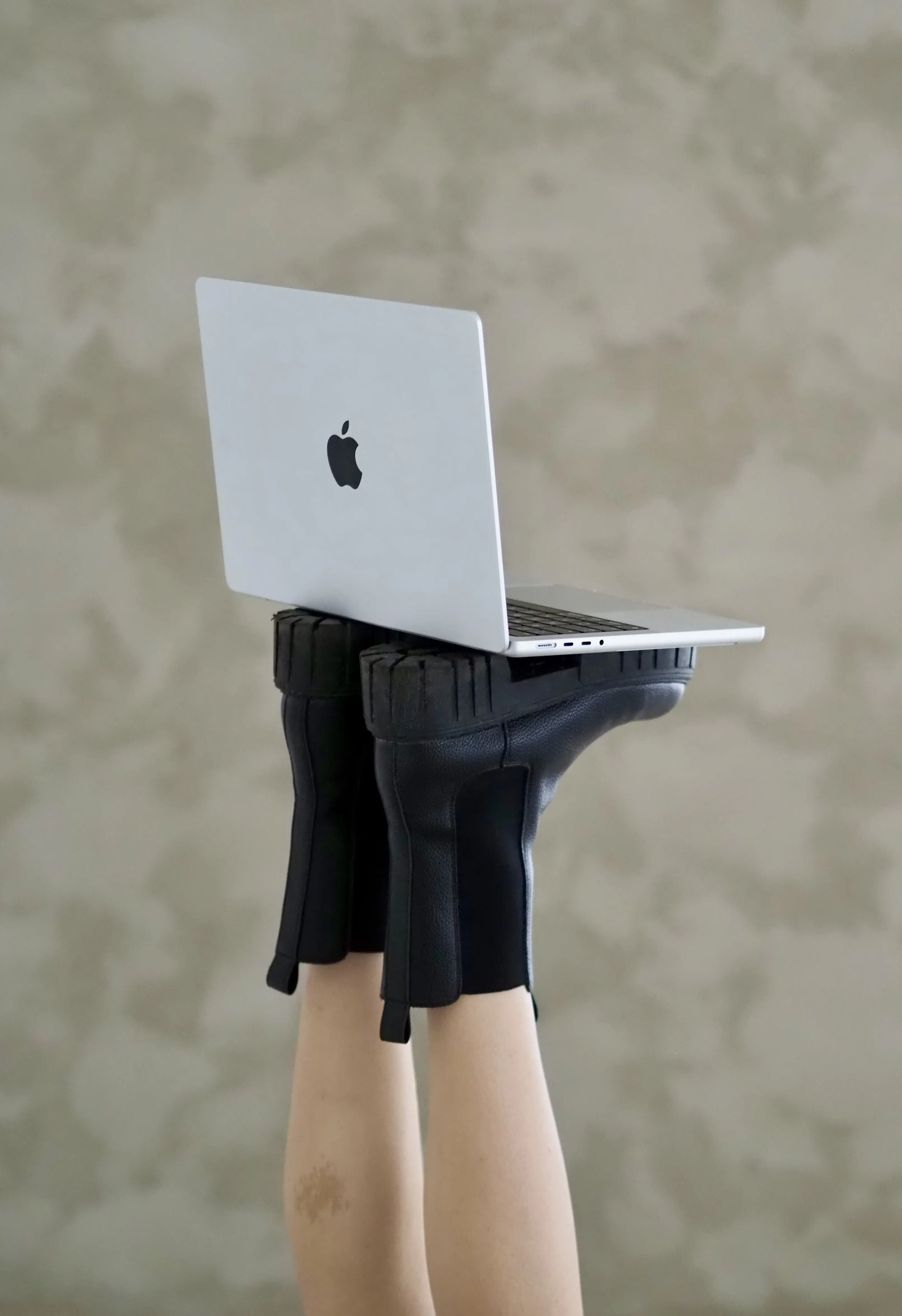 Ein silbernes MacBook ruht auf zwei schwarzen Stiefeln, die von einer Person getragen werden, deren Beine unten aus dem Bildrahmen verschwinden.