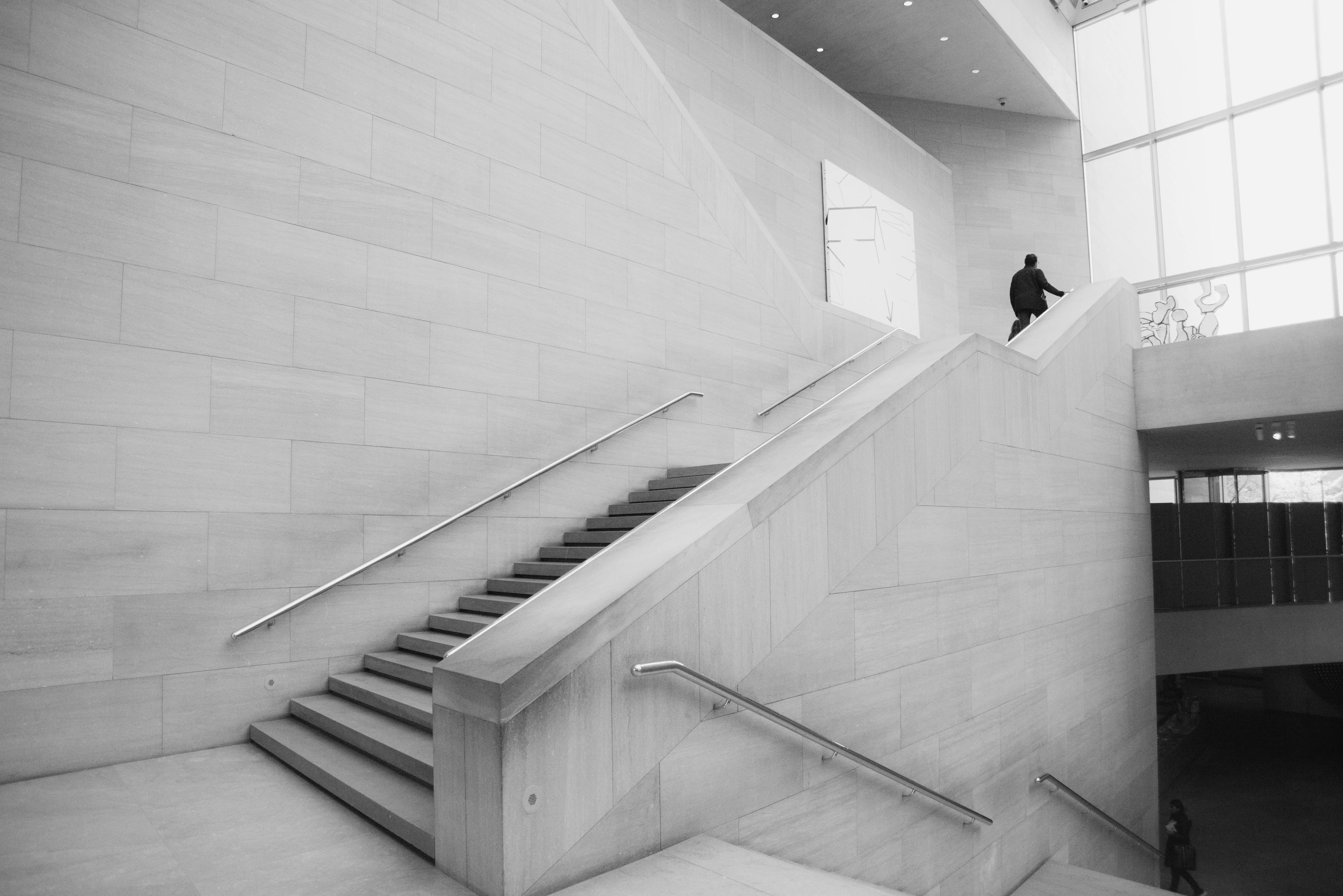 Ein minimalistisches Treppenhaus mit modernen Wänden aus hellem Stein. Eine Person geht die Treppe hinauf in Richtung eines großen Fensters mit viel natürlichem Licht.