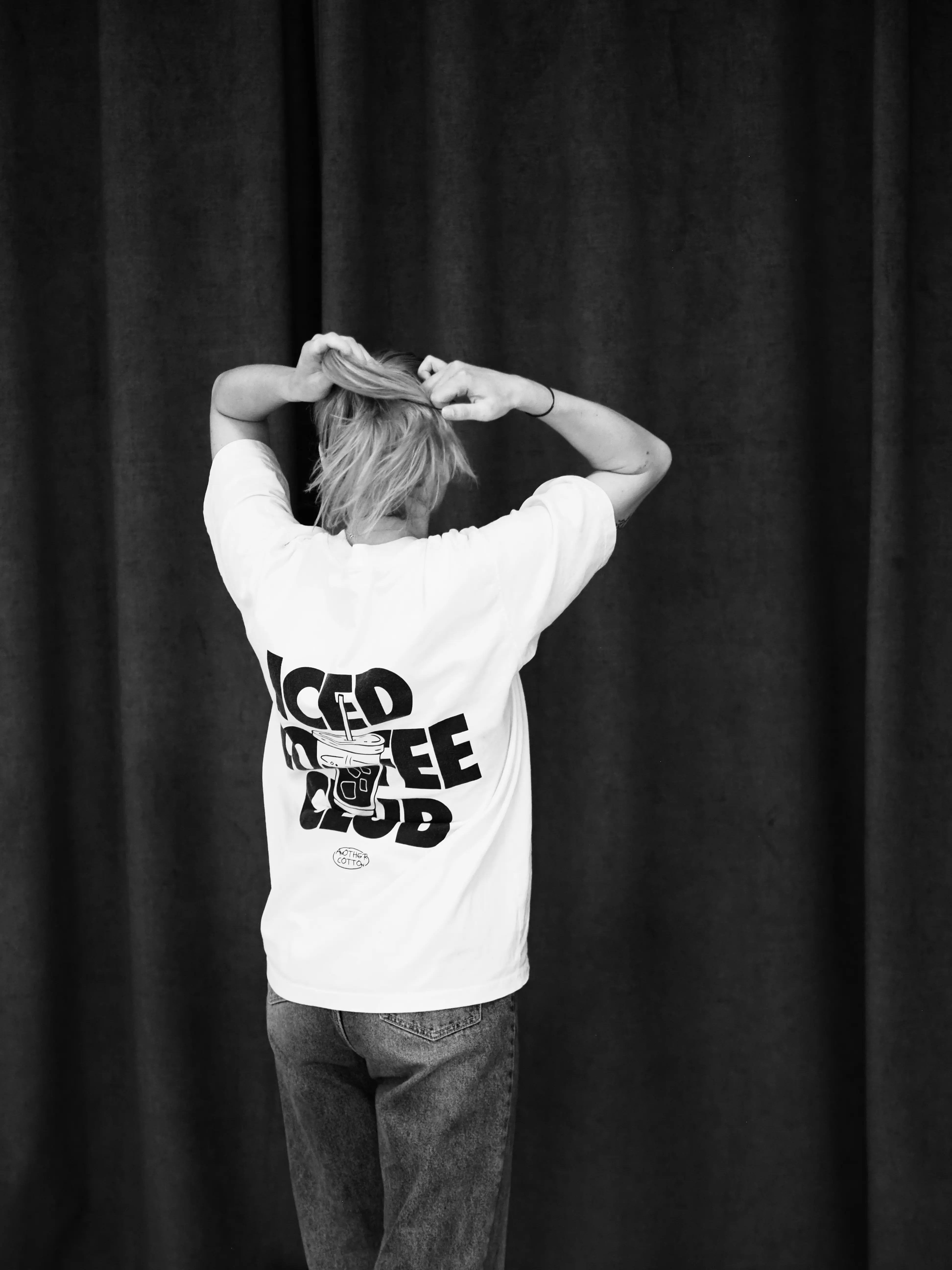 Eine Person steht mit dem Rücken zur Kamera vor einem dunklen Vorhang. Sie trägt ein weißes T-Shirt mit der Aufschrift 'Iced Coffee Club' und einem illustrierten Kaffeebecher auf der Rückseite. Die Person bindet sich die Haare zusammen.