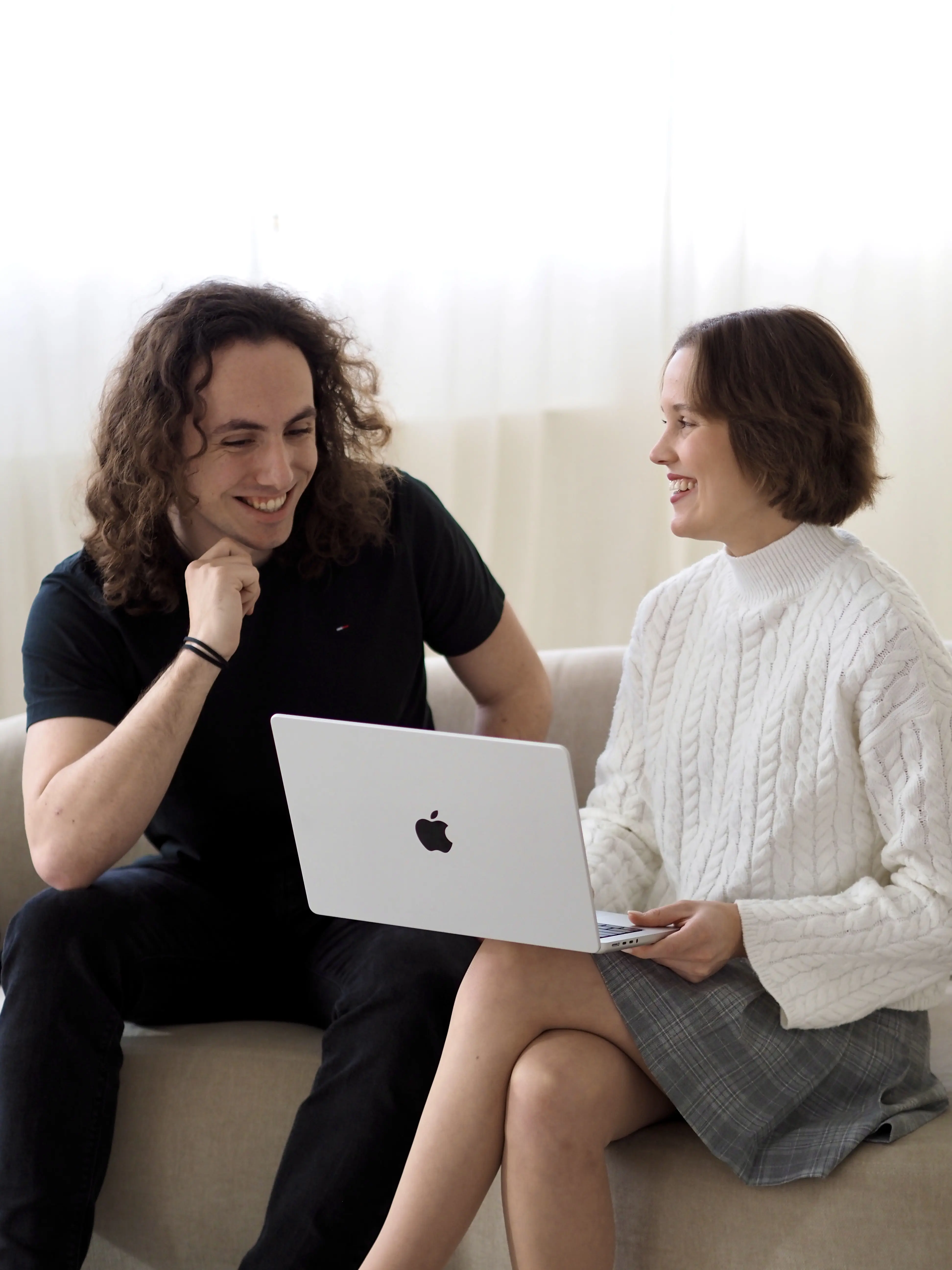 Zwei Personen sitzen zusammen auf einem Sofa und schauen lächelnd auf einen silbernen Laptop, den eine der Personen hält. Die Person links trägt ein schwarzes T-Shirt, die Person rechts trägt einen weißen Strickpullover und einen karierten Rock. Beide wirken entspannt und befinden sich in einer Kommunikation auf Augenhöhe