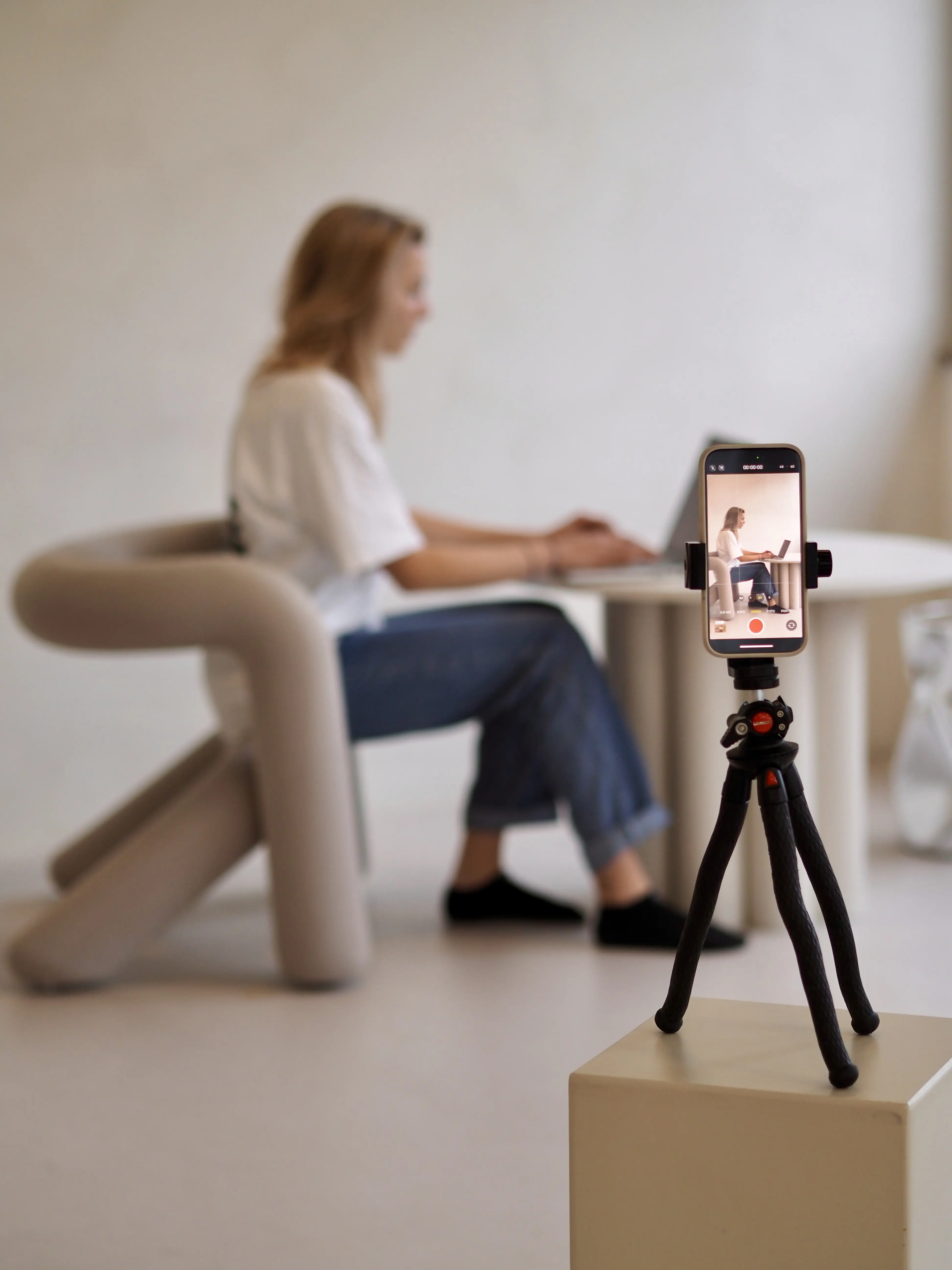 Im Vordergrund ist ein Smartphone auf einem Stativ zu sehen, das eine Aufnahme macht. Im Hintergrund sitzt eine Person in einem modernen Stuhl und arbeitet an einem Laptop. Die Szene vermittelt Professionalität und modernes Arbeiten.