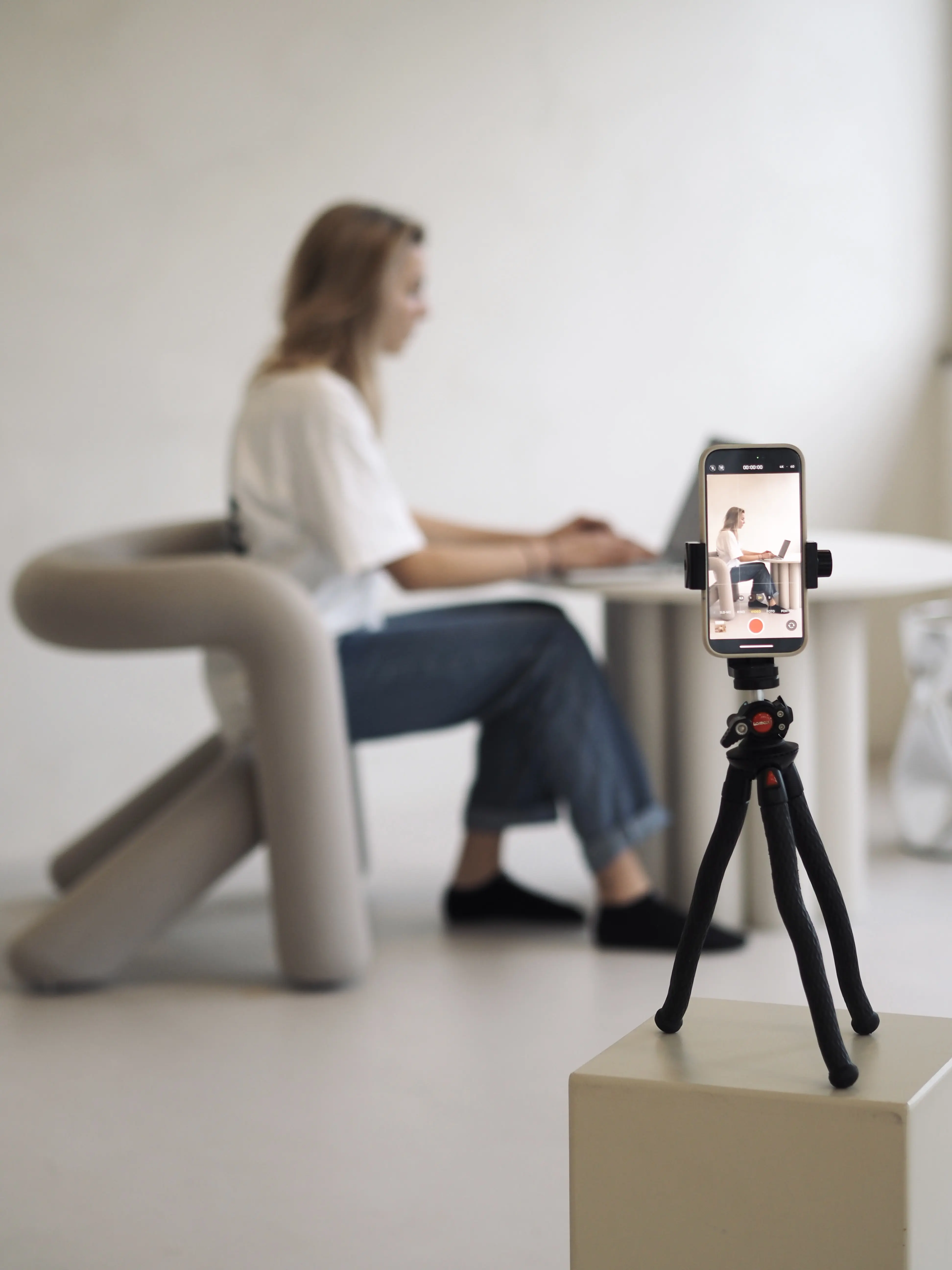 Eine Person sitzt im Hintergrund an einem Laptop und arbeitet an einem Projekt. Im Vordergrund ist ein Smartphone zu sehen, das in ein Stativ eingespannt ist, vermutlich für die Content-Erstellung.